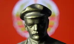 Figurka wodza w czapce, symbol totalitaryzmu, na tle czerwonej flagi z rozmazanym symbolem.