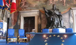 Dwa niebieskie krzesła z gwiazdami Unii Europejskiej, przed nimi stół z tym samym symbolem. W tle posąg papieża i malowidła.