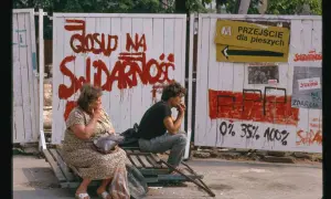 Dwie osoby siedzą na ławce przed płotem z napisem "Głosuj na Solidarność". To symboliczny obraz upadku komunizmu.