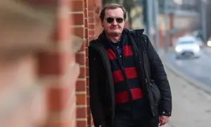 Mężczyzna w okularach przeciwsłonecznych, oparty o ceglaną ścianę, w zimowej kurtce i swetrze w pasy. Człowiek istota pełna sprzeczności, jak ten kontrast między surową ścianą a miejskim zgiełkiem w tle.
