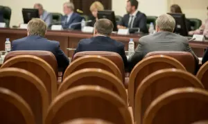 Mężczyźni w garniturach siedzą przy stole, czekając na **absolutorium**. W tle widać monitory i innych uczestników spotkania.