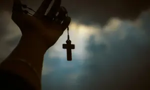 Dłoń trzymająca różaniec z krzyżykiem na tle zachmurzonego nieba.