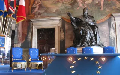 Dwa niebieskie krzesła z gwiazdami Unii Europejskiej, przed nimi stół z tym samym symbolem. W tle posąg papieża i malowidła.