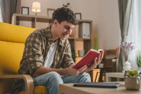 Uśmiechnięty młodzieniec w kratę, siedząc w żółtym fotelu, niedawno czy nie dawno pochłonął się lekturą czerwonej książki.
