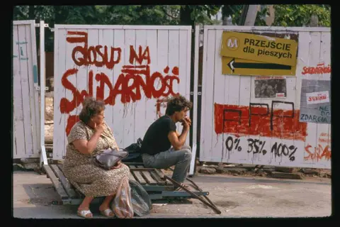 Dwie osoby siedzą na ławce przed płotem z napisem "Głosuj na Solidarność". To symboliczny obraz upadku komunizmu.