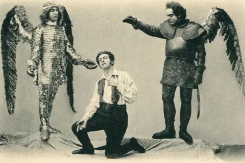 Trzech mężczyzn w strojach teatralnych: dwóch w skrzydłach i zbrojach, jeden klęczy.