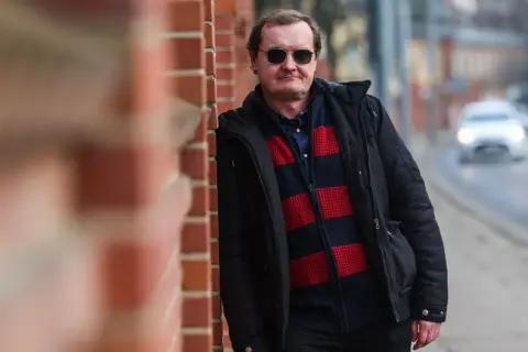 Mężczyzna w okularach przeciwsłonecznych, oparty o ceglaną ścianę, w zimowej kurtce i swetrze w pasy. Człowiek istota pełna sprzeczności, jak ten kontrast między surową ścianą a miejskim zgiełkiem w tle.