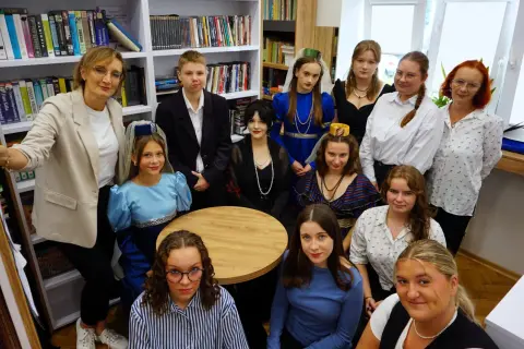 Grupa osób w strojach historycznych, jakby z powieści Dostojewskiego, pozuje na tle regałów z książkami.