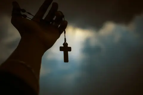 Dłoń trzymająca różaniec z krzyżykiem na tle zachmurzonego nieba.
