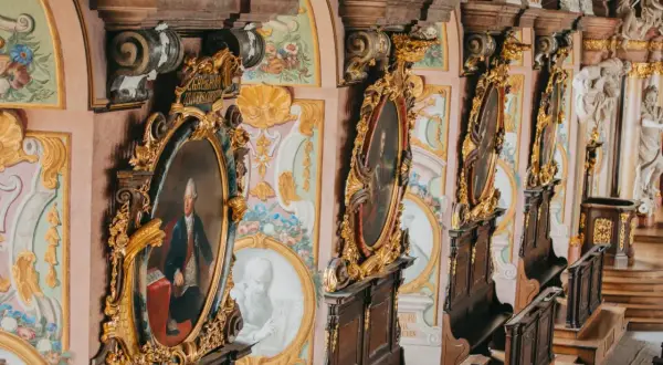 Wnętrze z portretami w złotych ramach, ozdobnymi malowidłami i rzeźbami. Idealizm epoki widoczny w kunsztownych detalach.