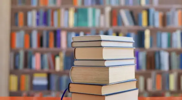Stos książek na biurku, tło biblioteki z kolorowymi grzbietami. Idealne lektury na e8.