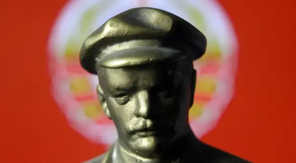 Figurka wodza w czapce, symbol totalitaryzmu, na tle czerwonej flagi z rozmazanym symbolem.