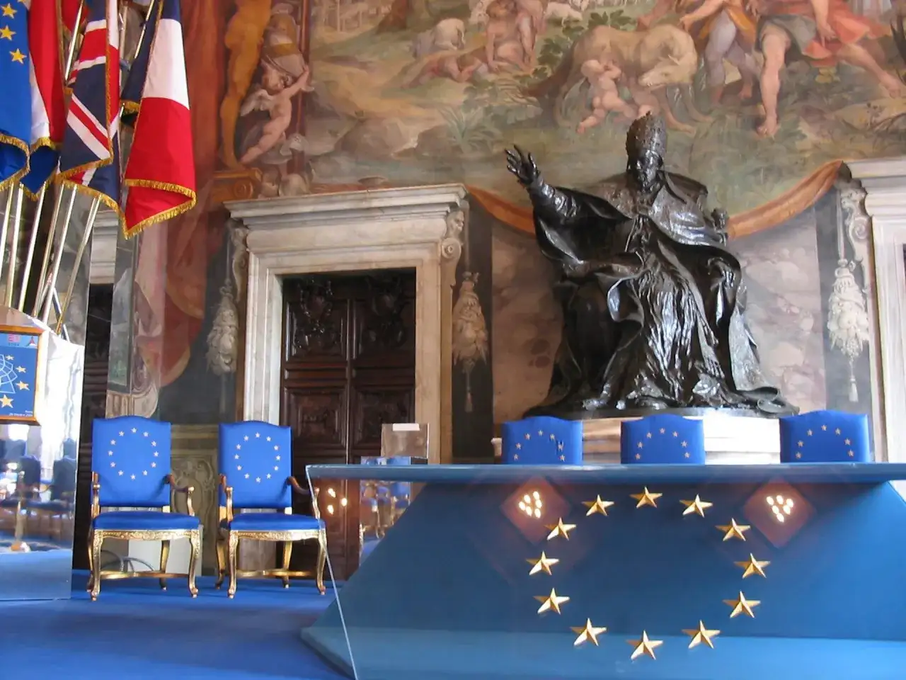 Dwa niebieskie krzesła z gwiazdami Unii Europejskiej, przed nimi stół z tym samym symbolem. W tle posąg papieża i malowidła.