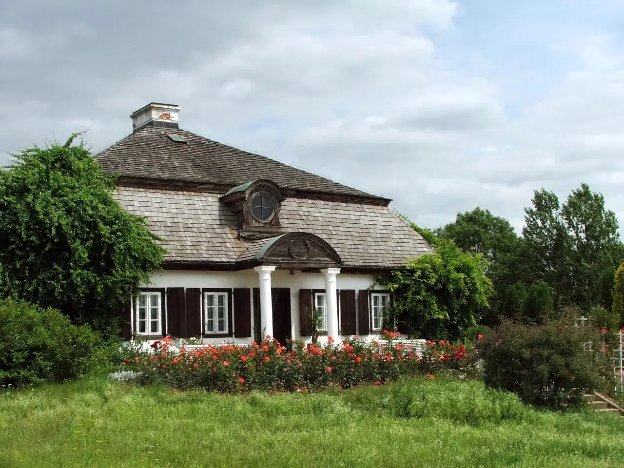 Dworek z werandą i kolumnami, otoczony kwitnącymi różami i zielenią. Scena jak z