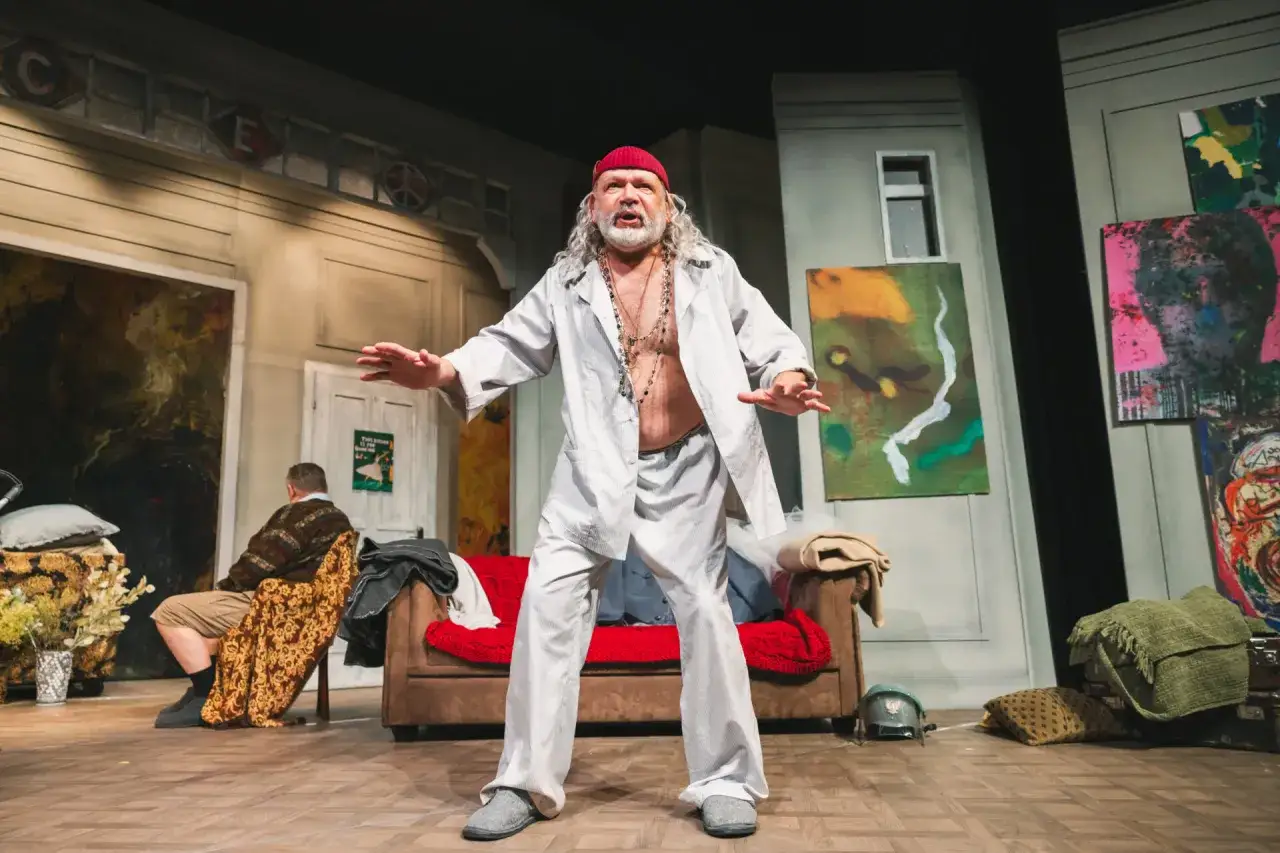 Scena teatralna z mężczyzną w czerwonej czapce i białej koszuli, gestykulującym w dziwaczny sposób. To groteska w tangu, pełna surrealistycznych obrazów i nonsensu.