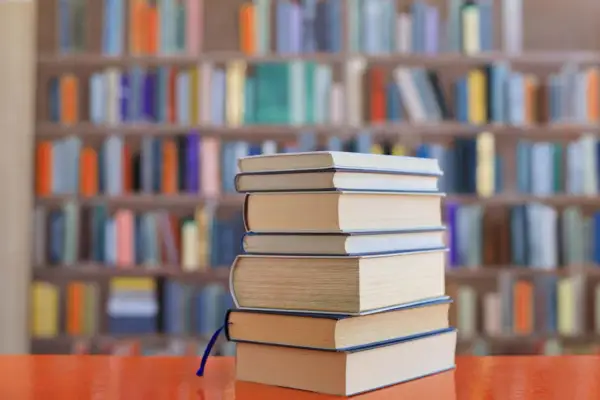 Stos książek na biurku, tło biblioteki z kolorowymi grzbietami. Idealne lektury na e8.