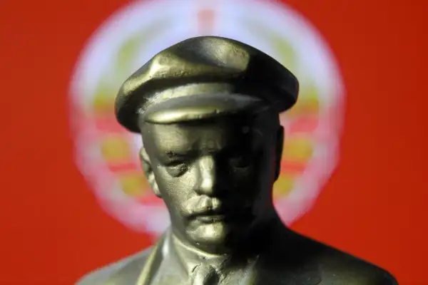 Figurka wodza w czapce, symbol totalitaryzmu, na tle czerwonej flagi z rozmazanym symbolem.