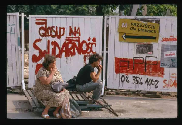 Dwie osoby siedzą na ławce przed płotem z napisem "Głosuj na Solidarność". To symboliczny obraz upadku komunizmu.