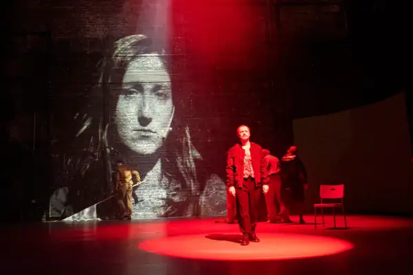 Na scenie, w świetle czerwonych reflektorów, stoi mężczyzna. W tle projekcja przedstawia twarz matki Cezarego Baryki.