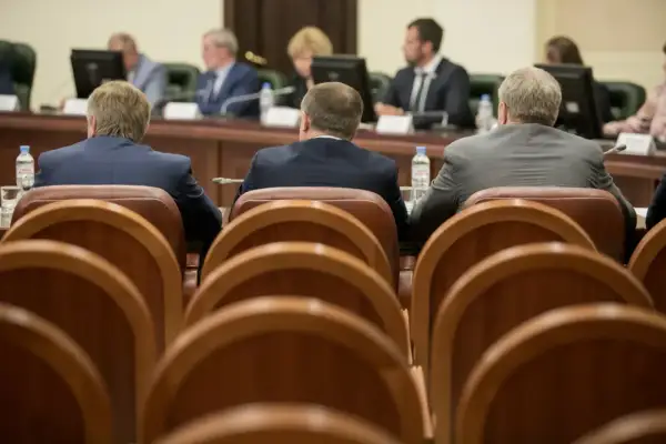 Mężczyźni w garniturach siedzą przy stole, czekając na **absolutorium**. W tle widać monitory i innych uczestników spotkania.